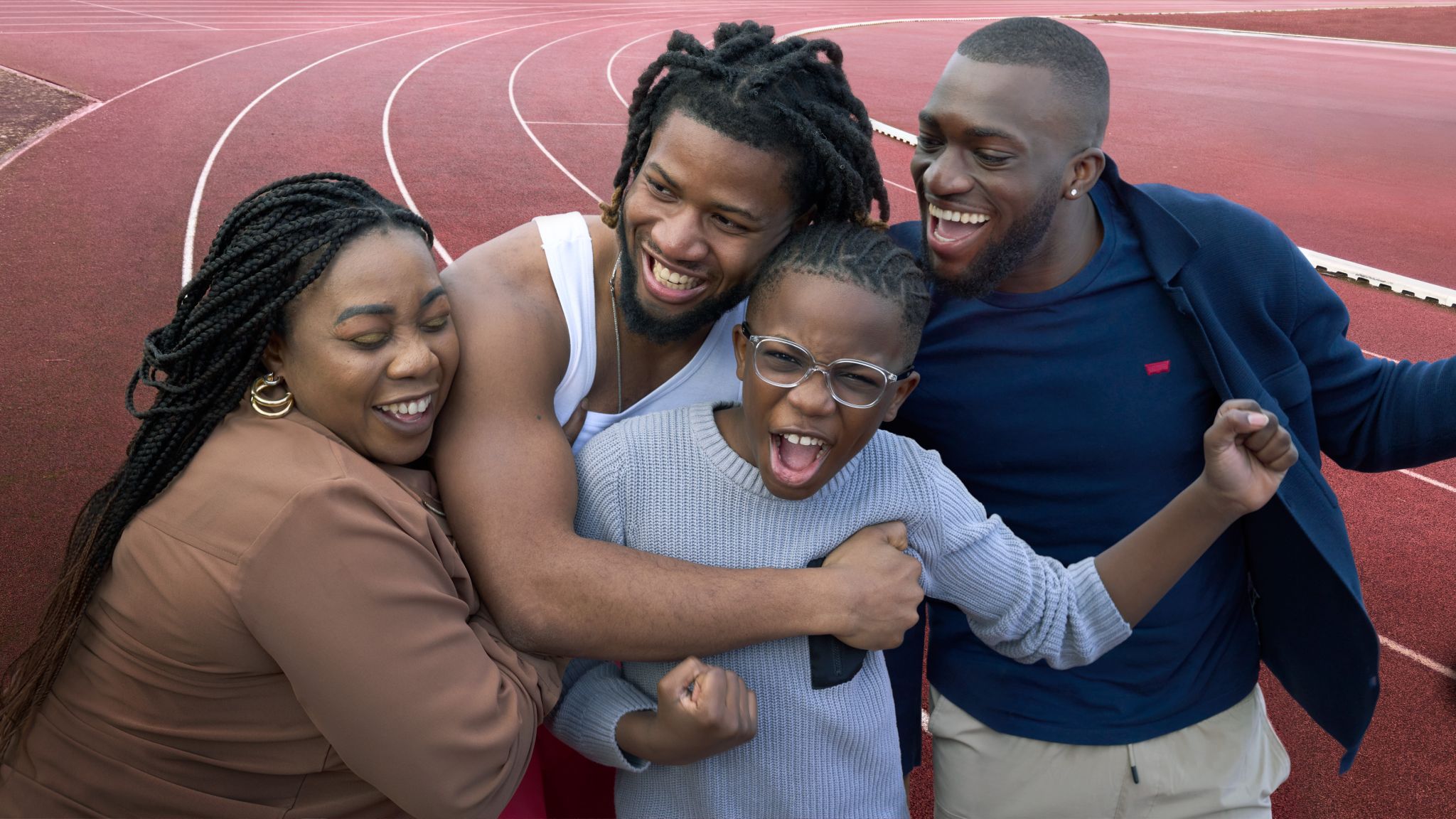 A smiling family group at a running track. An adult woman embraces Para athlete Emmanuel Oyinbo-Coker in a white tank top. They are joined by a laughing boy with glasses and another smiling man in a blue shirt, all sharing a happy moment together.